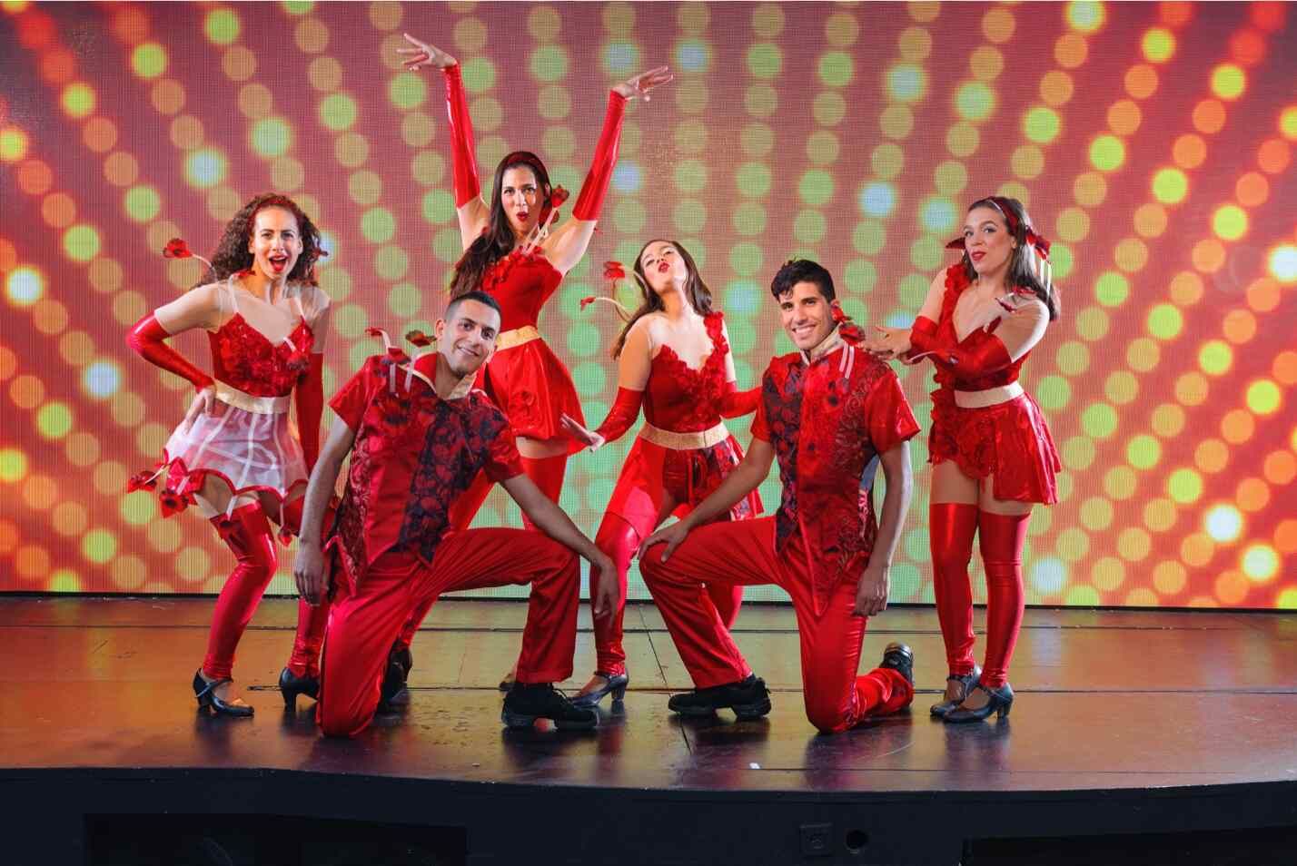 קבוצת שישה רקדנים ורקדניות לבושים בתלבושות בולטות באדום עומדת על במה עם רקע צבעוני מנוקד