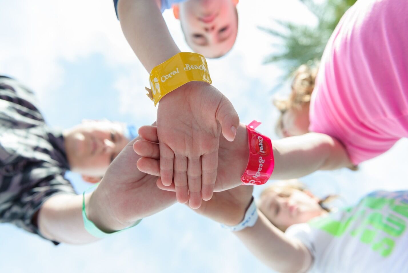 קבוצת ילדים מצמידה ידיים כאשר על פרקי ידיהם צמידים צבעוניים עם הכיתוב  Coral Beach