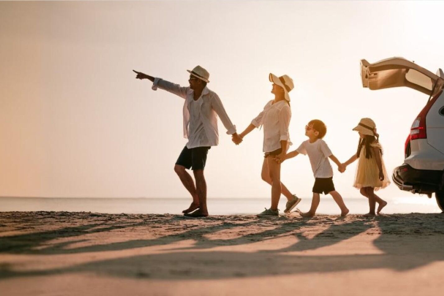 משפחה יוצאת לטיול חוף בשקיעה, ההורים מחזיקים ידיים עם ילדיהם ליד רכב עם תא מטען פתוח