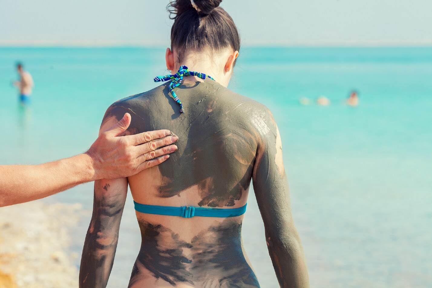 גב של אישה בחוף ים המלח מרוחה בבוץ מינרלי, בזמן שיד עוזרת לה למרוח את הבוץ על גב